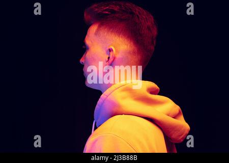Profilansicht eines jungen Mannes mit Hoodie isoliert über dunklem Studiohintergrund in Neonlicht. Konzept der Emotionen, Mode Stockfoto
