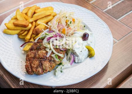 Bifteki, griechisches Hackfleischsteak gefüllt mit Feta-Käse, serviert mit pommes frites, Krautsalat, Tsatziki und Zwiebeln auf einem weißen Teller, hölzernes Outdoor-Ta Stockfoto