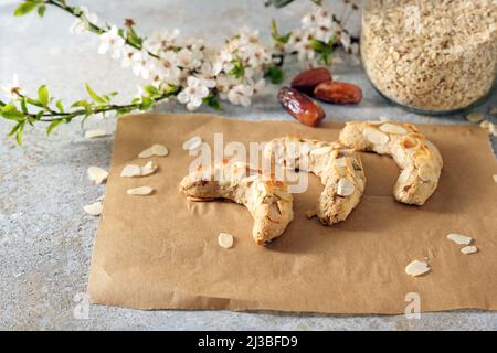 Gesunde festliche Kekse aus Mandeln, Haferflocken und Datteln auf Backpapier, Zutaten und einem blühenden Frühlingszweig auf rustikalem grauen Hintergrund, Cop Stockfoto