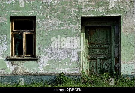 Eine alte Holztür und ein kaputtes Fenster mit einem verfallenen antiken dekorativen weißen Holzrahmen, die an der Wand des Familienhauses angebracht sind. Stockfoto