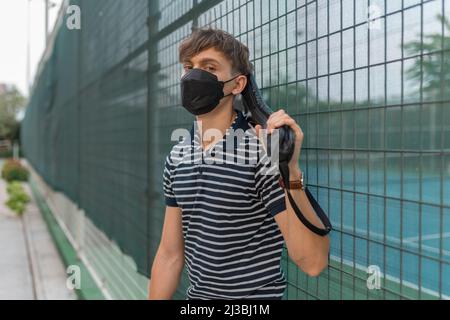 Junger Mann mit schwarzer Gesichtsmaske, der auf dem Tennisplatz steht und einen Tennisschläger hält. Hochwertige Fotos Stockfoto