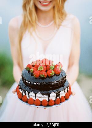 Hochzeit Schokoladenkuchen Nahaufnahme mit Erdbeeren in den Händen der Smimimimg Braut in rosigen Hochzeitskleid. Rustikale Hochzeit im Freien. Stockfoto