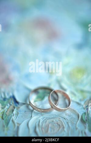Klassische Eheringe auf blauem Hintergrund mit Platz für Inschrift, flaches Lay, Draufsicht. Weicher abstrakter Blumenhintergrund mit bemalten Rosen. Weichfoc Stockfoto
