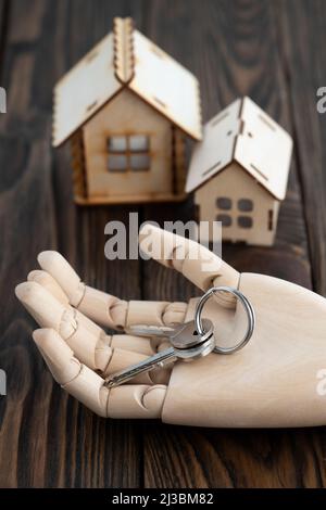 Hölzerne Puppenhand mit Schlüsseln auf der Handfläche mit Spielzeug-Holzhäusern auf einem Hintergrund auf einem Tisch Stockfoto