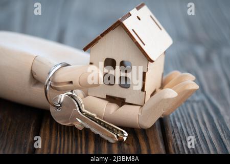 Puppenhand aus Holz mit einem hölzernen Spielzeughaus auf der Handfläche mit Schlüsseln auf dem Tisch Stockfoto