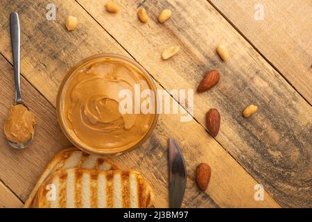 Schüsseln mit Erdnussbutter und Erdnüssen auf dunklem Holzboden aus der Sicht von oben, reichhaltiges Frühstück Stockfoto