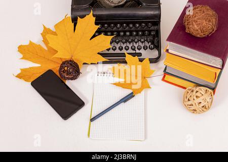 Herbststimmung. Auf dem Tisch stehen alte Schreibmaschine, modernes Mobiltelefon, Bücher, Notizbuch und Stift, trockene gelbe Ahornblätter. Arbeitsplatz des Studenten oder Schriftstellers. Stockfoto