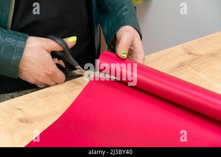 Nahaufnahme der Hände einer Floristin, die mit einer Schere gefärbtes, leuchtend rosa Geschenkpapier für einen Blumenstrauß in einem Blumenladen schneidet Stockfoto