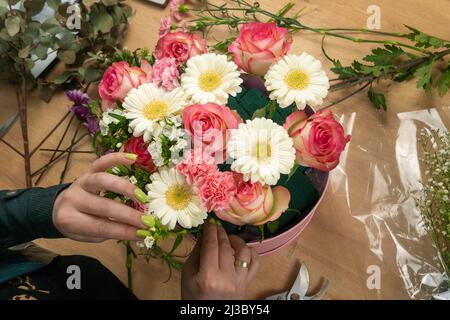 Draufsicht auf die Hände einer jungen Floristin, die eine wunderschöne Komposition aus zarten rosa Rosen und weißen Gänseblümchen auf dem Tisch kreiert Stockfoto