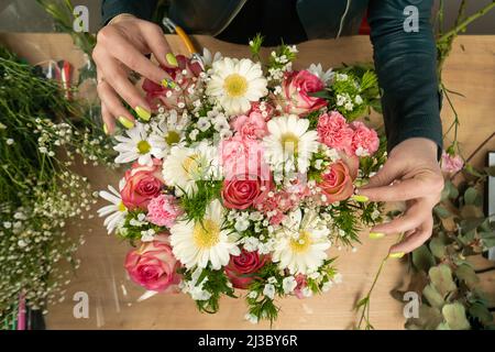 Draufsicht auf die Hände einer jungen Floristin, die eine wunderschöne Komposition aus zarten rosa Rosen, Nelken und weißen Gänseblümchen auf dem Tisch kreiert Stockfoto