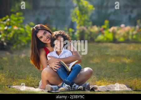 Mutter und Sohn schauten zusammen auf die Kamera, während sie mit einem Buch im Park saßen Stockfoto