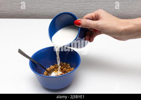 Gießt Milch aus einem Becher mit der Hand in einen Teller mit Müsli Stockfoto