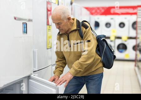 Älterer grauhaariger Rentner, der am Schalter im Ausstellungsraum der Abteilung für Elektrogeräte einen Kühlschrank sah Stockfoto