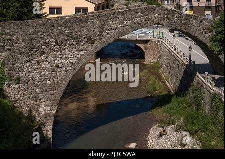 Ein Fliegenfischer in Wadern wirft seine Leine, während er auf dem schnell fließenden Fluss Llobregat in La Pobla de Lillet in Katalonien, Spanien, zwischen einer modernen Straßenbrücke und einer alten Mautbrücke, dem anmutigen, einbeinigen Bogen Pont Vell, steht, der in den 1300s v. Chr. erbaut wurde. Die Pont Vell hatte vielleicht einmal einen zweiten Bogen und die Aschenpappe ihrer Säulen deutet darauf hin, dass ihre Ursprünge römisch sein könnten. Stockfoto