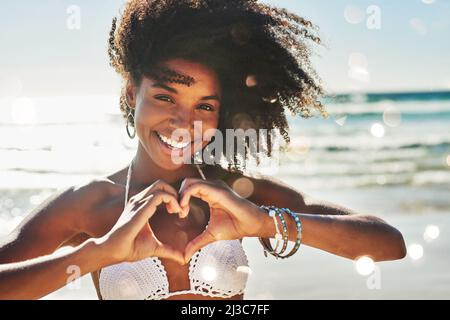 Mein Herz gehört zum Sommer. Porträt einer schönen jungen Frau, die mit ihren Händen am Strand eine herzförmige Geste macht. Stockfoto