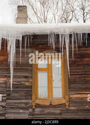 Schmelzende transparente Eiszapfen hängen am Rand des Dachs. Vor dem Hintergrund der Holzwand des alten Hauses. Große Kaskaden, sogar schöne Reihen. Wolkiger Wintertag, weiches Licht. Stockfoto