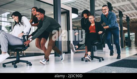 Fröhliche Kollegen genießen die gemeinsame Spielzeit bei der Arbeit. Fröhliche Gruppe von Kollegen, die sich in einem modernen Büro auf Stühlen schieben. Team von businesspeop Stockfoto
