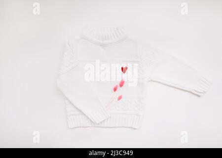 Weißer Wollpullover mit einem roten herzförmigen Pin auf weißem Hintergrund Draufsicht. Liebe, Romantik, Modekonzept. Flach liegend, Kopierplatz Stockfoto