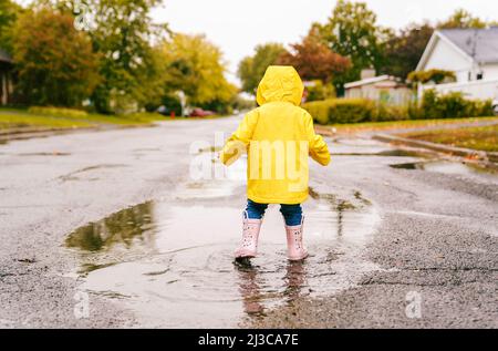 Lustige niedliche Baby-Mädchen tragen gelben wasserdichten Mantel und Stiefel spielen im Regen Stockfoto