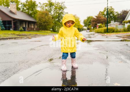 Lustige niedliche Baby-Mädchen tragen gelben wasserdichten Mantel und Stiefel spielen im Regen Stockfoto