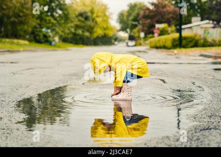 Lustige niedliche Baby-Mädchen tragen gelben wasserdichten Mantel und Stiefel spielen im Regen Stockfoto