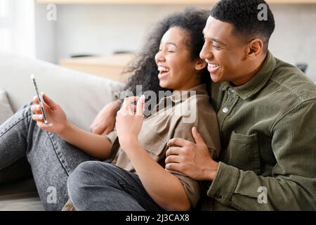 Seitenansicht des verliebten Paares sitzen zu Hause auf der Couch und nutzen Smartphone für Online-Gespräche, Video-Treffen mit Freunden Stockfoto