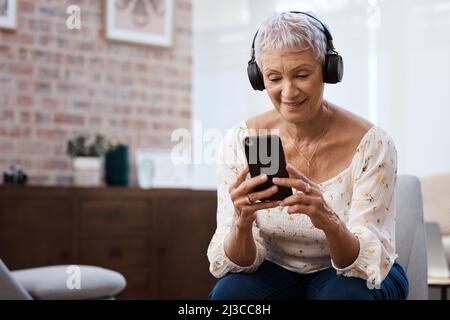 Eine neue Art, alte Lieder zu hören. Aufnahme einer älteren Frau mit Smartphone und Kopfhörer auf dem Sofa zu Hause. Stockfoto