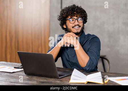Schöner junger Freiberufler mit Bart in einer Brille, der am Tisch sitzt und wegschaut, eine Pause von der Arbeit am Laptop macht, vom Urlaub träumt, von zu Hause aus arbeitet Stockfoto