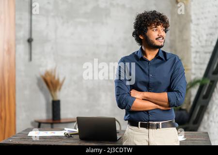 Indischer Geschäftsmann, der mit einem Laptop in einem modernen Büro neben dem Schreibtisch steht und wegschaut. Ehrgeiziger, lächelnder, multiethnischer Mann in eleganter Freizeitkleidung mit gekreuzten Armen Stockfoto