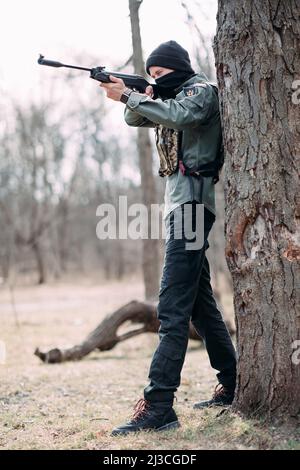 Der junge Mann übt und zielt von der in Körperpanzerung und Sturmhaube im Wald getragene Pumppistole ab. Stockfoto