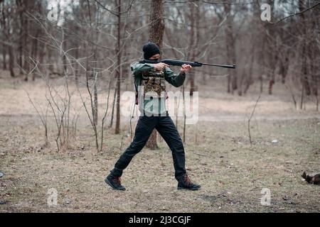 Der junge Mann übt und zielt von der in Körperpanzerung und Sturmhaube im Wald getragene Pumppistole ab. Stockfoto