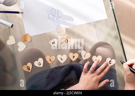 Draufsicht auf Crop anonymous Kid Writing No Mobbing Inschrift auf kleinen Herzen, während sie am Glastisch im Zimmer sitzen Stockfoto