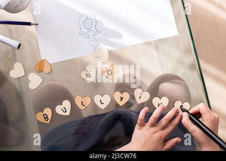 Draufsicht auf Crop anonymous Kid Writing No Mobbing Inschrift auf kleinen Herzen, während sie am Glastisch im Zimmer sitzen Stockfoto