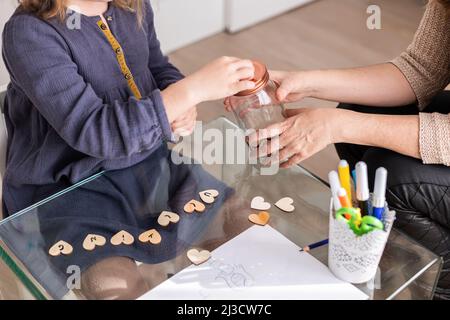 Cropped unkenntlich Mädchen machen Wunsch mit Glas kleinen hölzernen Herzen, während in der Nähe weibliche Psychologin sitzen Stockfoto