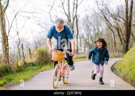 Ganzer Körper des aktiven Großvaters Fahrrad der Enkelin in der Nähe positiv laufenden Mädchen auf dem Weg in der Landschaft mit grünen Bäumen reiten Stockfoto