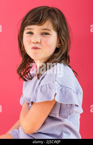 Seitenansicht von niedlichen kleinen Mädchen mit braunen Haaren machen Grimasse und Blick auf die Kamera vor rosa Hintergrund Stockfoto