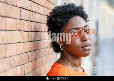 Seitenansicht einer unemotionalen afroamerikanischen Frau in farbenfroher Sonnenbrille, die mit einem Lächeln auf die Kamera blickt, während sie in der Nähe eines Gebäudes auf der Straße der Stadt steht Stockfoto