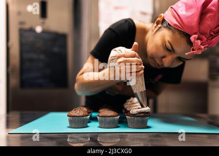 Hispanische Bäckerin im Kopftuch mit einem Gebäckbeutel, um während der Arbeit in der Backstube die Creme auf Schokoladenkuchenstücke zu quetschen Stockfoto