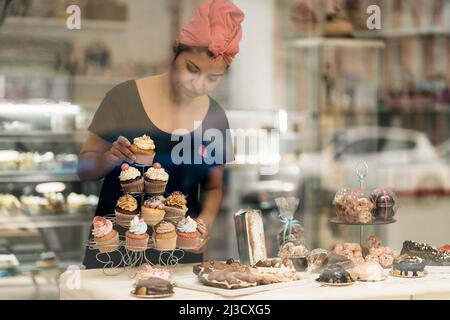 Hispanische Frau im Kopftuch, die tagsüber in der Bäckerei in der Nähe des Fensters Desserts auf dem Tisch arrangiert Stockfoto