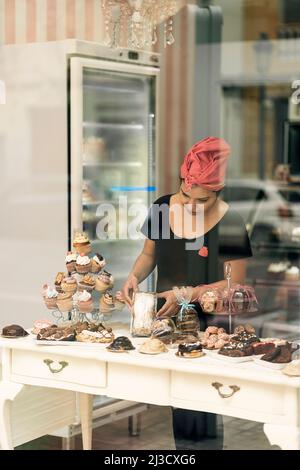Hispanische Frau im Kopftuch, die tagsüber in der Bäckerei in der Nähe des Fensters Desserts auf dem Tisch arrangiert Stockfoto