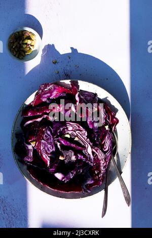 Blick von oben auf frischen veganen Salat aus reifem Rotkohl, der tagsüber in einer Schüssel mit Geschirr in der Nähe von Kapern auf einem sonnenbeschienenen Tisch serviert wird Stockfoto
