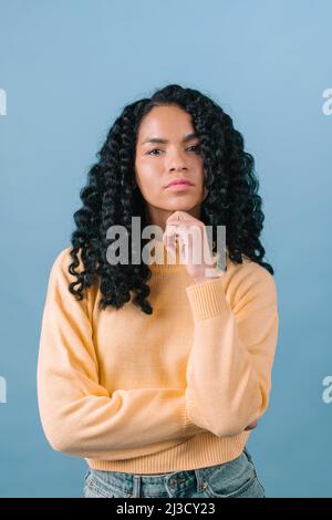 Nachdenkliche afroamerikanische Frau mit schwarzen lockigen Haaren und Blick auf die Kamera, während sie im hellen Studio auf blauem Hintergrund steht Stockfoto