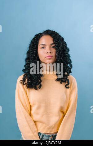Nachdenkliche afroamerikanische Frau mit schwarzen lockigen Haaren und Blick auf die Kamera, während sie im hellen Studio auf blauem Hintergrund steht Stockfoto
