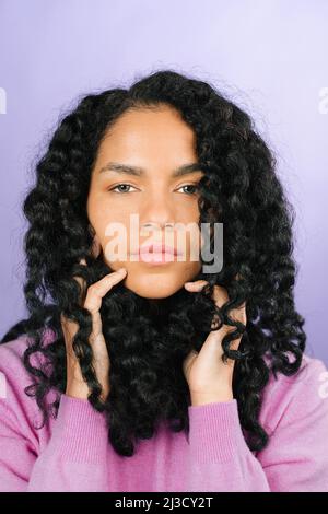 Nachdenkliche afroamerikanische Frau mit schwarzen lockigen Haaren und Blick auf die Kamera, während sie im hellen Studio auf violettem Hintergrund steht Stockfoto