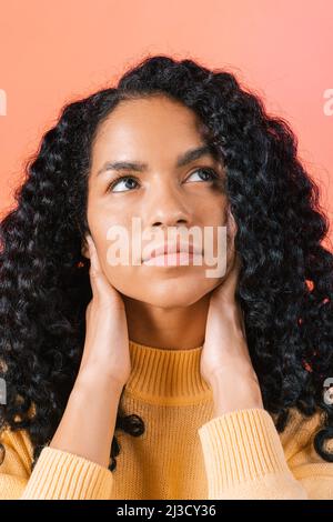 Nachdenkliche afroamerikanische Frau mit schwarzen lockigen Haaren und wegschauen, während sie im hellen Studio auf orangefarbenem Hintergrund steht Stockfoto