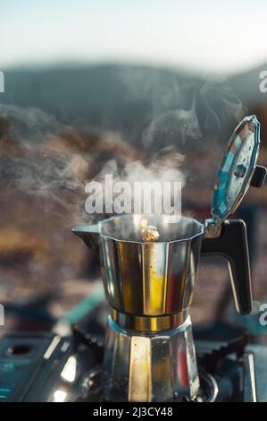 Metalltopf mit geöffnetem Deckel auf dem tragbaren Gasherd zum Brauen von frischem aromatischem Kaffee auf dem Campingplatz Stockfoto