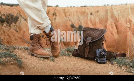 Nicht erkennbarer Mann in weißen Hosen und Lederschuhen, der in der Nähe von Wildlederrucksack und Fotokamera gegen den unartigen Canyon in der Natur steht Stockfoto