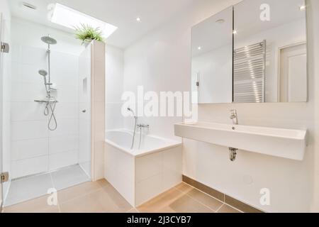 Weißes Keramikwaschbecken an der Wand mit Spiegel in der Nähe der Badewanne im hellen, geräumigen Badezimmer mit Duschkabine und gefliesten Wänden Stockfoto