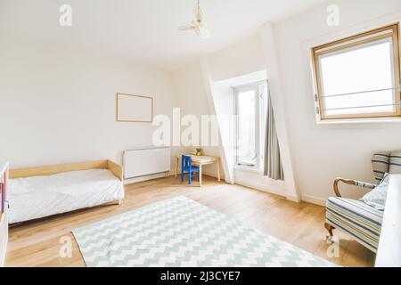 Weißes Bett in der Nähe von einem kleinen Tisch in einem geräumigen, hellen Kinderschlafzimmer mit Teppich und Fenstern in einem Apartment mit bequemem Sessel Stockfoto