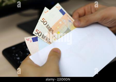 Briefumschlag mit Euro-Banknoten in männlichen Händen. Mann zieht Geld aus einem Umschlag auf PC-Tastatur Hintergrund, Löhne, Bonus oder Bestechung Konzept Stockfoto